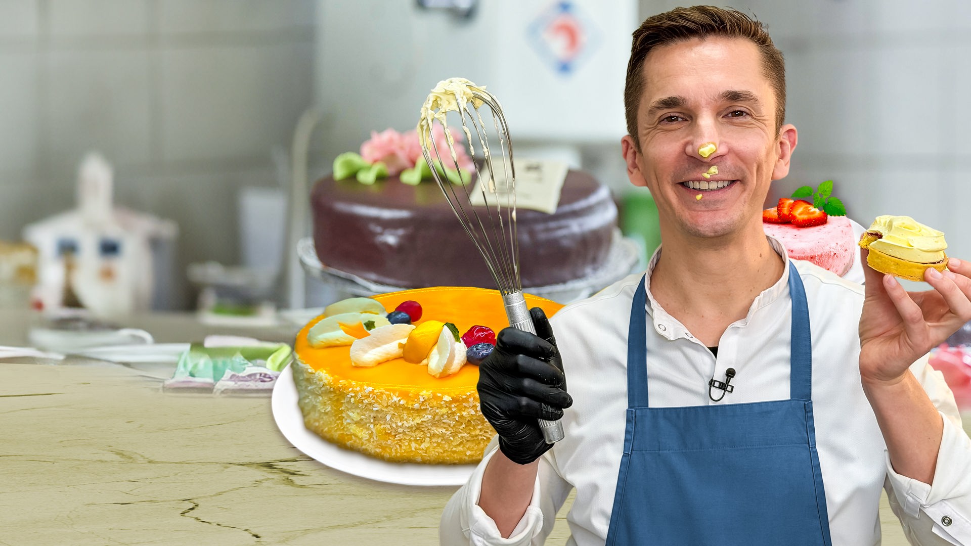 Eric Mayer steht in einer Küche und schaut freudig in die Kamera. Er trägt ein weißes Hemd und darüber eine blaue Schürze. In der rechten Hand hält er einen mit Creme verschmierten Schneebesen. In der linken Hand hält er ein Mini-Törtchen. Auf seiner Nase und rund um den Mund sind Kleckse aus Tortencreme. Um ihn herum stehen auf einer Küchenarbeitsplatte drei verschiedene Torten platziert.