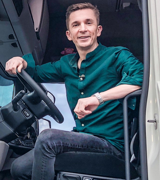 Moderator Eric Mayer sitzt im Fahrerhaus eines weiß-blauen LKW. Die Tür steht offen.