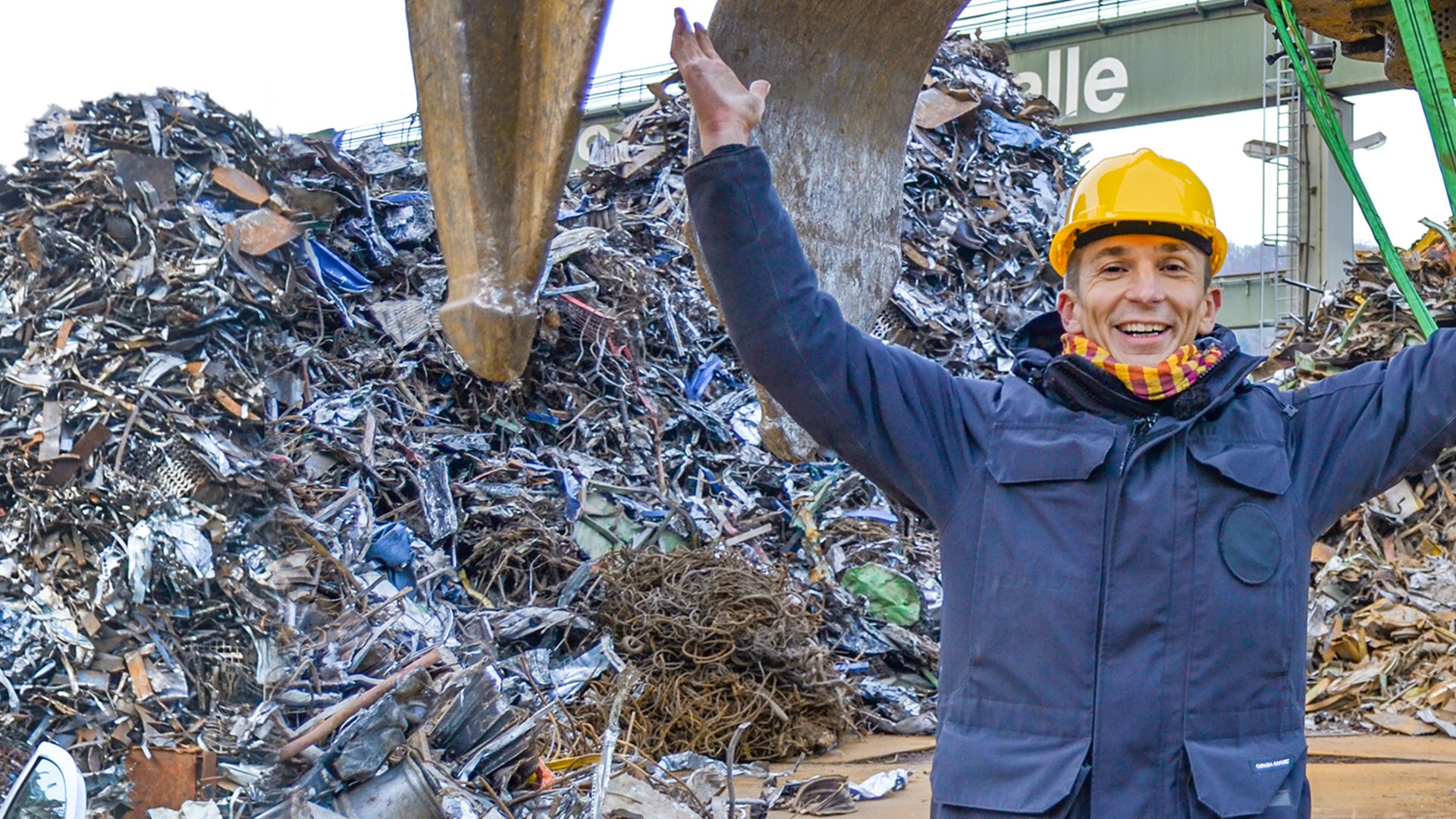PUR+ Moderator Eric Mayer steht auf einem Schrottplatz inmitten von Bergen aus Metallschrott. Hinter ihm befindet sich der riesige Greifarm eines Krans. Daran ist eine Apparat befestigt, der messen kann, welches Gewicht allein mittels Magnetismus gehoben werden kann.