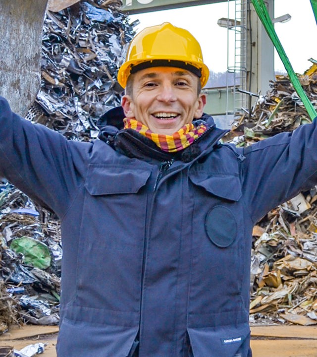 PUR+ Moderator Eric Mayer steht auf einem Schrottplatz inmitten von Bergen aus Metallschrott. Hinter ihm befindet sich der riesige Greifarm eines Krans. Daran ist eine Apparat befestigt, der messen kann, welches Gewicht allein mittels Magnetismus gehoben werden kann.