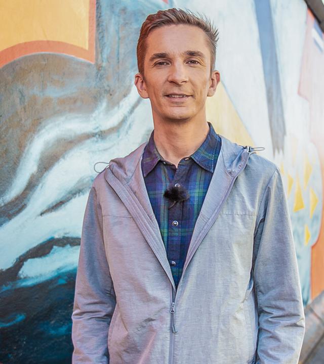 PUR+ Moderator Eric Mayer steht vor einem erhalten gebliebenen Stück der Berliner Mauer und schaut freundlich in die Kamera. Im Hintergrund sind weitere Menchen zu sehen, die sich die Mauer anschauen.