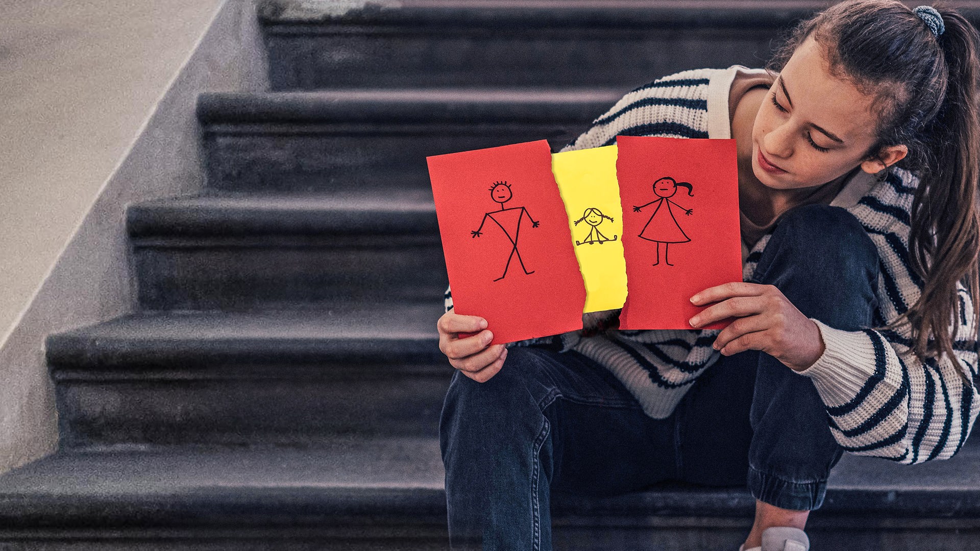 Eine jugendliches Mädchen sitzt auf einer Treppe. In den Händen hält sie zwei rote Karten, dazwischen eine gelbe Karte. Auf den roten Karten ist ein Mal ein weibliches Strichmännchen und ein Mal ein männliches Strichmännchen abgebildet. Die gelbe Karte zeigt eine Strichzeichnung eines kleinen Mädchens.