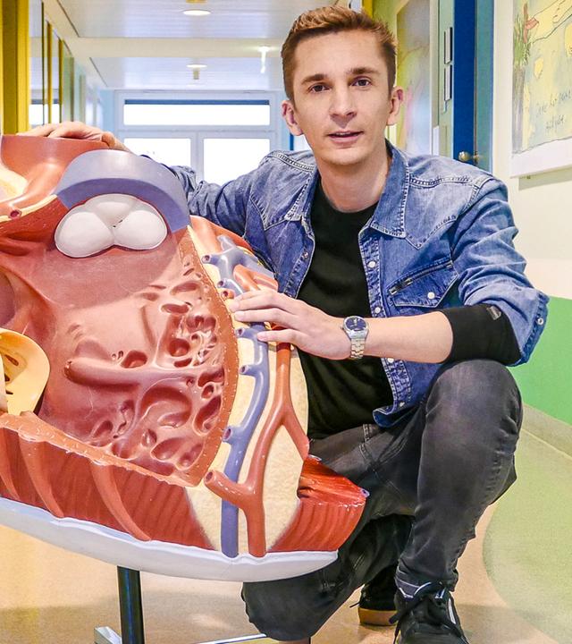 PUR+-Moderator Eric Mayer kniet neben einem überdimensionalen 3-D-Modell einer Herzkammer auf einem Krankenhausflur.