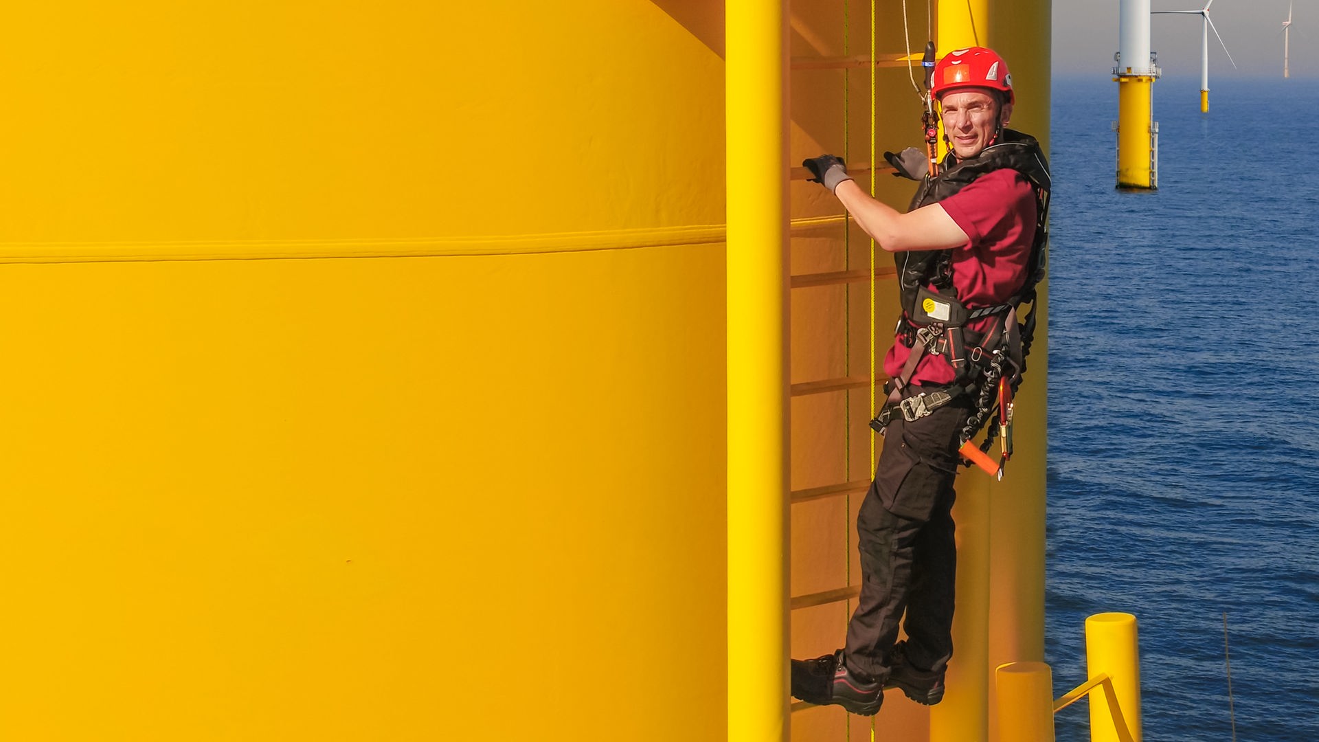 Moderator Eric Mayer klettert an der gelben Außenwand eines Windrads hoch. Er ist ausgesattet mit Klettergeschirr und Helm und gut gesichert. Im Hintergrund stehen weitere Windräder in der Nordsee.