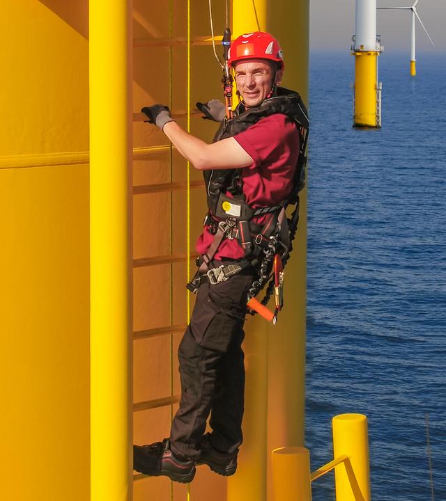Moderator Eric Mayer klettert an der gelben Außenwand eines Windrads hoch. Er ist ausgesattet mit Klettergeschirr und Helm und gut gesichert. Im Hintergrund stehen weitere Windräder in der Nordsee.