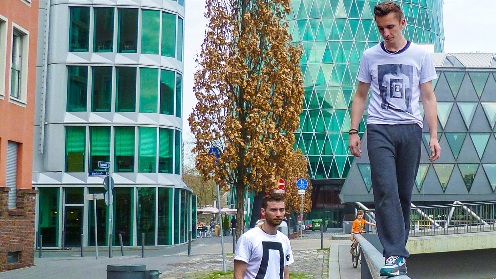 PUR+ Moderator Eric Mayer im urbanen Umfeld umgeben von Bürogebäuden mit glitzernden Fassaden. Er balanciert vorsichtig auf einem Handlauf.
