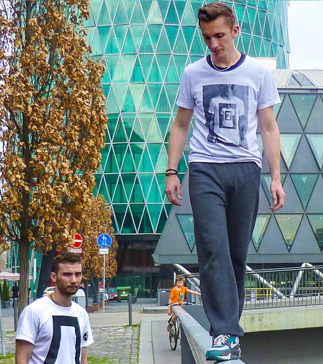 PUR+ Moderator Eric Mayer im urbanen Umfeld umgeben von Bürogebäuden mit glitzernden Fassaden. Er balanciert vorsichtig auf einem Handlauf.