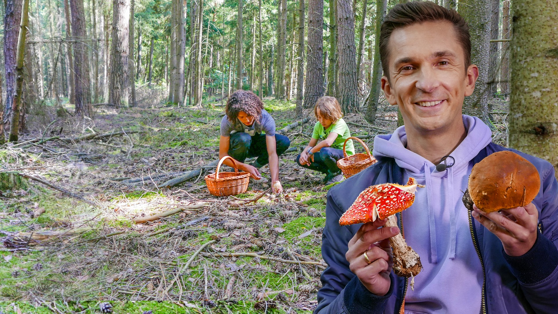 Eric Mayer steht im Wald zwischen den Bäumen und direkt vor einem Baumstamm. Er lächelt in die Kamera. In seiner rechten Hand hält er einen großen Fliegenpilz. In der linken Hand hält er einen großen Steinpilz. Im Hintergrund suchen ein Kind und eine erwachsene Person hockend am Waldboden nach Pilzen. Sie haben einen Korb dabei.