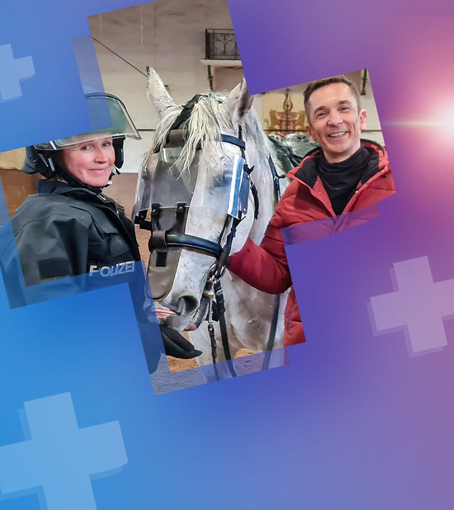 PUR+ - Eric steht zusammen mit einer Polizistin, die eine Uniform mit Helm trägt und einem Helm in einer Reithalle. Zwischen ihnen steht ein Polizeipferd, das für seinen Einsatz trainiert wird.