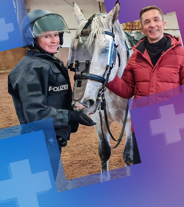 PUR+ - Eric steht zusammen mit einer Polizistin, die eine Uniform mit Helm trägt und einem Helm in einer Reithalle. Zwischen ihnen steht ein Polizeipferd, das für seinen Einsatz trainiert wird.