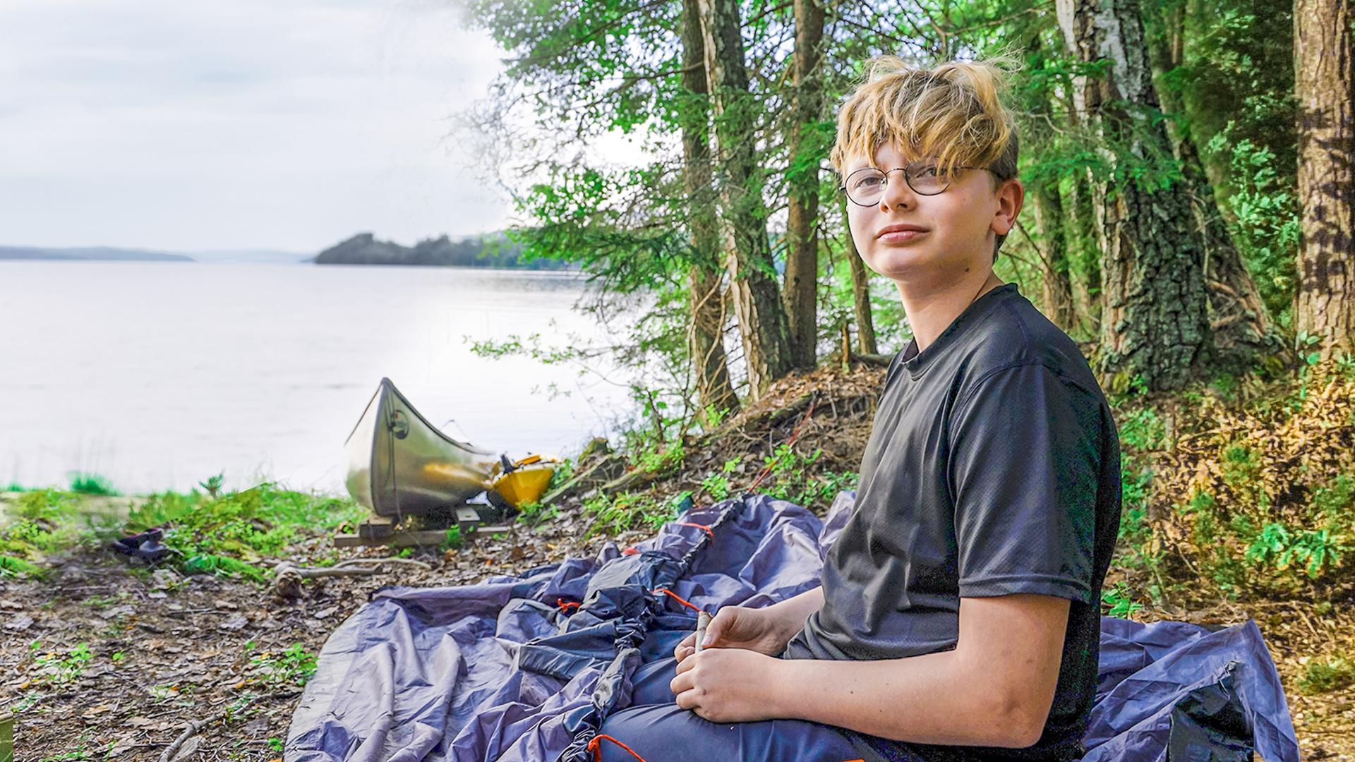 Marc sitzt auf einem steinigen Untergrund am Rande eines großen Sees. Neben ihm liegt das noch nicht aufgebaute Zelt. Ein paar Meter weiter liegt ein Kanadier am Ufer. Hinter dem See liegen bewaldete Berge.