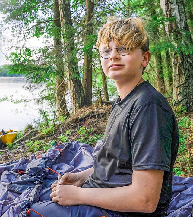 Marc sitzt auf einem steinigen Untergrund am Rande eines großen Sees. Neben ihm liegt das noch nicht aufgebaute Zelt. Ein paar Meter weiter liegt ein Kanadier am Ufer. Hinter dem See liegen bewaldete Berge.
