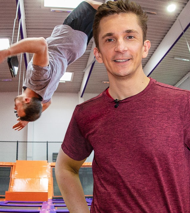 Eric Mayer steht im Vordergrund des Bildes, dahinter springt Amadei Weiland einen Rückwärtssalto in einer Trampolinhalle.