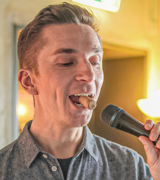 Eric Mayer steht in einem Tonstudio. Rechts von ihm ist ein Lautsprecher. Er hat einen Korken im Mund und hält eine Mikrofon mit seiner linken Hand vor den Mund.