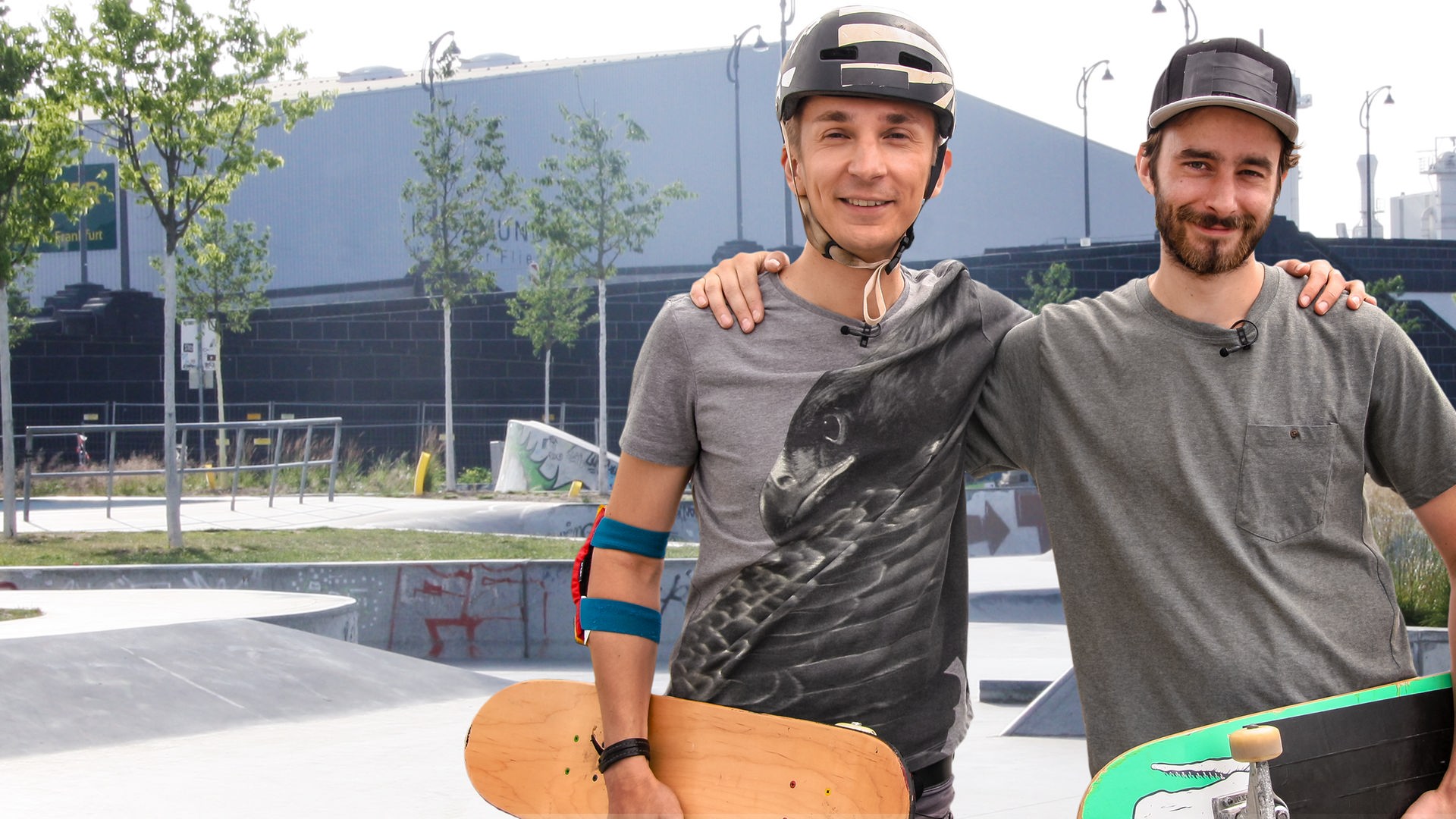 Eric Mayer und sein Trainer stehen auf einer Skateranlage. Sie halten ihre Boards im Arm. Eric trägt einen Schutzhelm und Ellbogenschützer. Der Trainer trägt eine Kappe. Beide schauen lächelnd nach vorne.