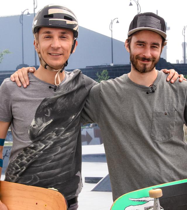 Eric Mayer und sein Trainer stehen auf einer Skateranlage. Sie halten ihre Boards im Arm. Eric trägt einen Schutzhelm und Ellbogenschützer. Der Trainer trägt eine Kappe. Beide schauen lächelnd nach vorne.