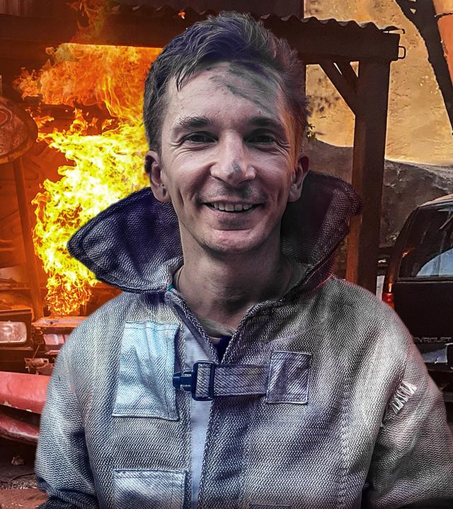 Moderator Eric Mayer trägt einen feuerfesten Spezialanzug, den er für eine gefährliche Stuntszene mit Feuer braucht. Im Hintergrund sieht man einen Sportwagen, aus dem eine Feuersäule in die Höhe steigt.