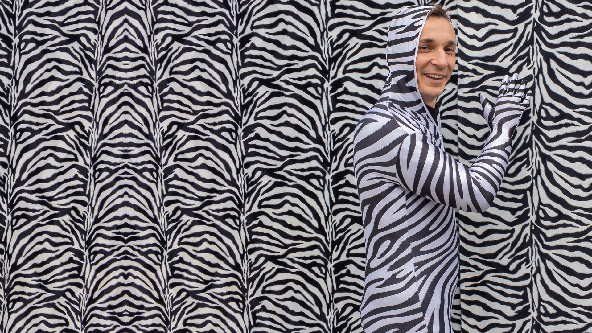 PUR+ Sendung Tarnung: Moderator Eric Mayer steht in einem Ganzkörper-Kostüm im Zebralook vor einer Wand im Zebralook. Nur sein Gesicht trägt keine Zebrastreifen und ist ungeschminkt.