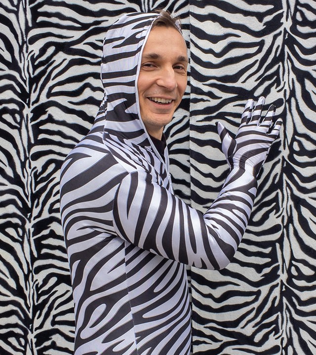 PUR+ Sendung Tarnung: Moderator Eric Mayer steht in einem Ganzkörper-Kostüm im Zebralook vor einer Wand im Zebralook. Nur sein Gesicht trägt keine Zebrastreifen und ist ungeschminkt.