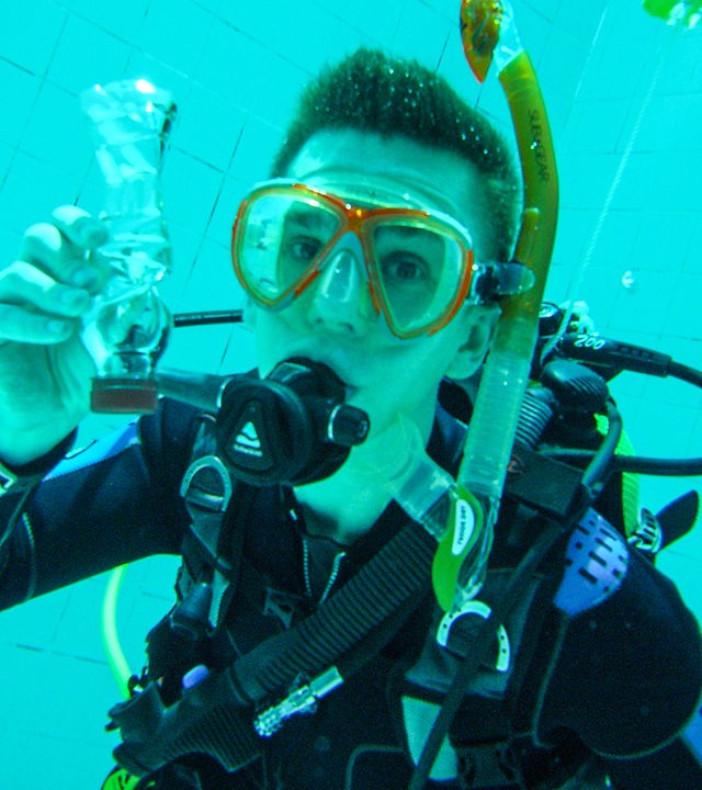 Moderator Eric Mayer im Taucheranzug und mit voller Tauchausrüstung unter Wasser in einem Schwimmbecken. Dieses ist der Drucktank der Uni Rostock. Er hat hält eine Plastikflasche in der Hand, die auf Grund des herrschenden Drucks stark zusammengedrückt ist.