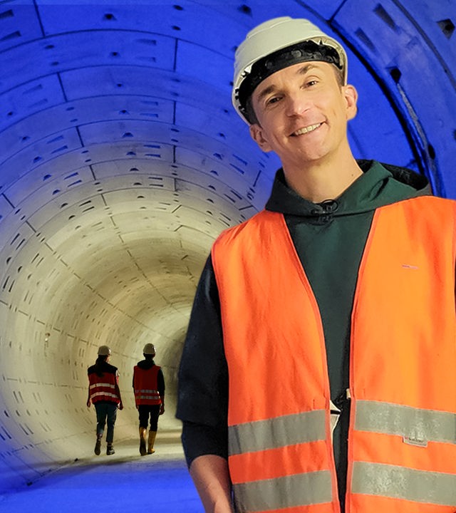 Moderator Eric Mayer steht rechts neben einem blau beleuchteten Tunnel aus Betonelementen. Er trägt eine orangene, neonfarbene Sicherheitsweste und einen Baustellenhelm. Im Tunnelhintergrund laufen zwei weitere Bauarbeitende.