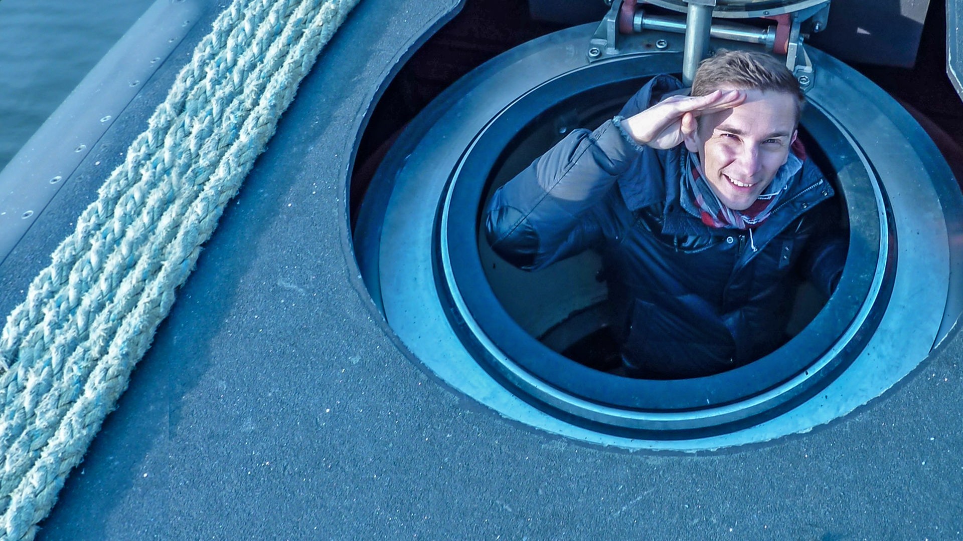 Moderator Eric Mayer ist auf einem U-Boot im Meer. Er schaut aus der geöffneten Luke heraus und grüßt mit der rechten Hand, indem er die Handkante an die Stirn hält.