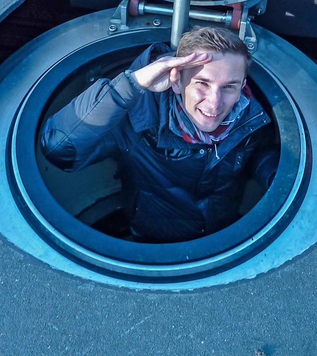 Moderator Eric Mayer ist auf einem U-Boot im Meer. Er schaut aus der geöffneten Luke heraus und grüßt mit der rechten Hand, indem er die Handkante an die Stirn hält.