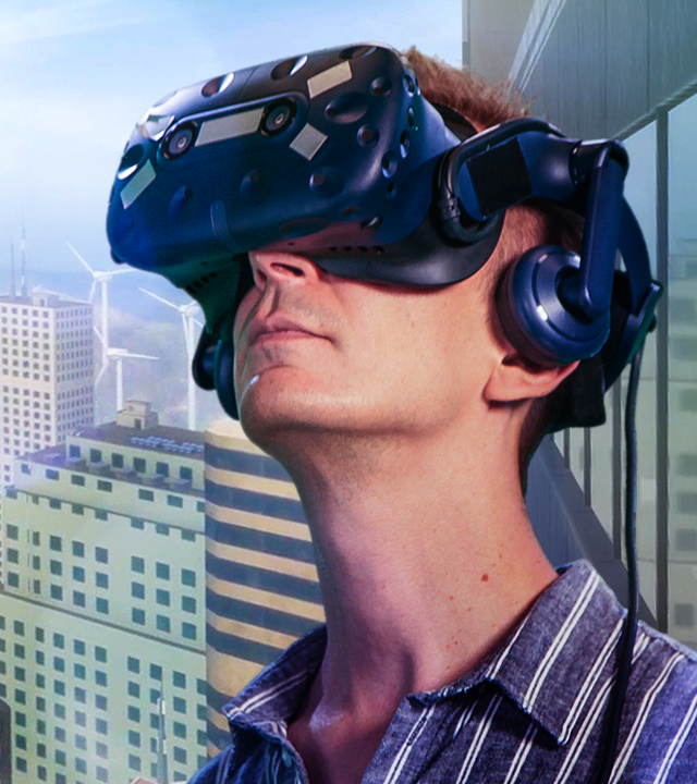 PUR+ Moderator Eric Mayer steht mit einer VR-Brille auf dem Kopf vor einer vom Computer erzeugten Stadtkulisse mit vielen Hochhäusern.