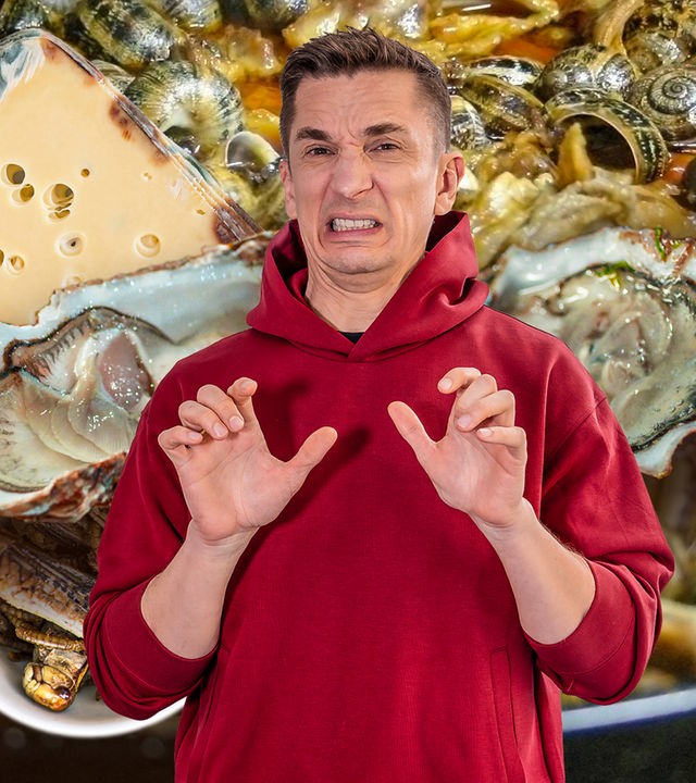 Eric Mayer steht in einem roten Kapuzenpullover mit Gesicht zur Kamera. Er schaut angewidert, verzieht Mund und Nase und macht mit den Händen eine abwehrende Geste. Im Hintergrund sind geöffnete Austern, schimmeliger Käse, gekochte Schnecken und frittierte Insekten zu sehen.