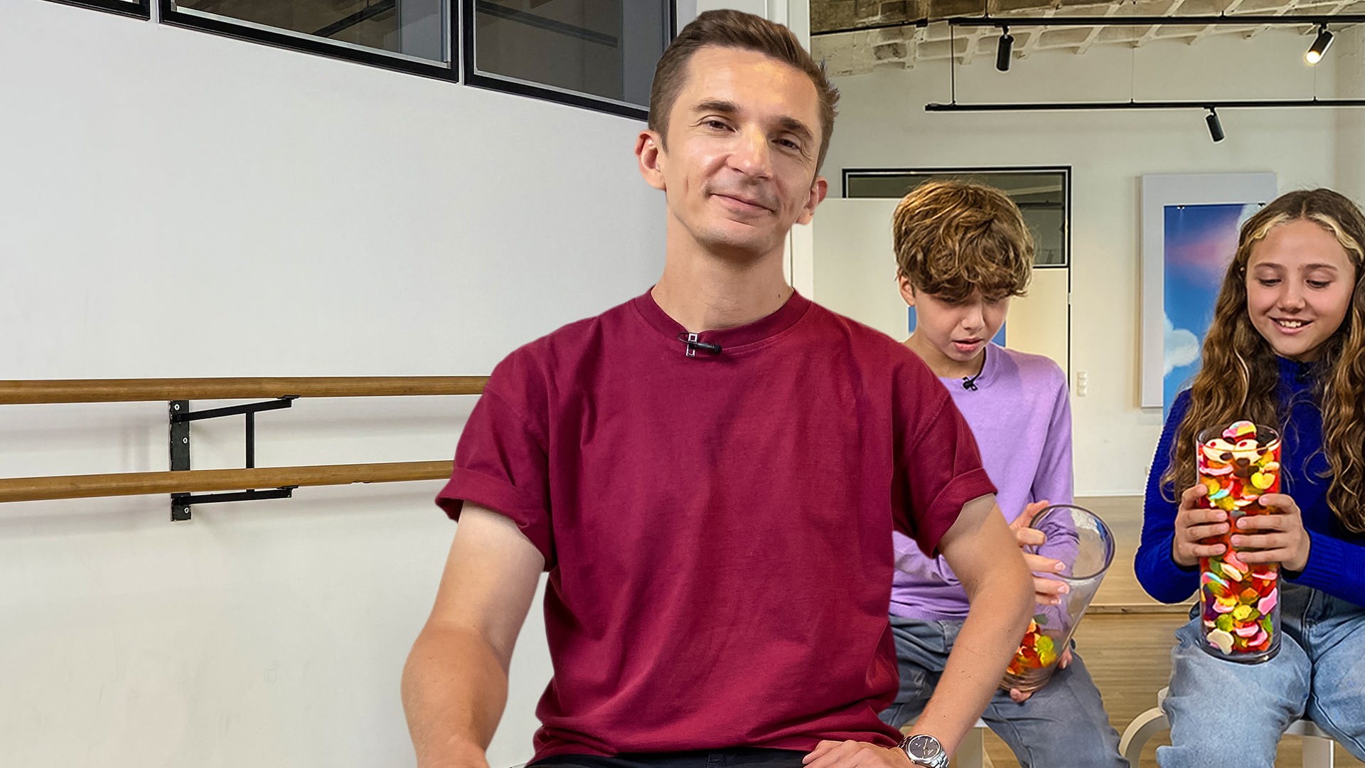 Moderator Eric Mayer steht in einem weißen Raum. Er trägt ein bordauxrotes Shirt und schaut in die Kamera. Hinter ihm sitzen zwei Jugendliche, ein Mädchen und ein Junge. Sie hält ein prall mit Süßigkeiten gefülltes, großes Glas in der Hand. Sein Glas ist nur ein bisschen mit Süßigkeiten gefüllt. Sie freut sich. Er schaut skeptisch. Im Hintergrund an der Wand hängt das PUR+-Logo. 