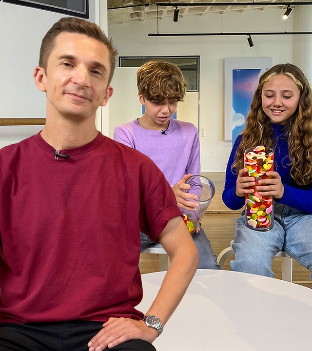 Moderator Eric Mayer steht in einem weißen Raum. Er trägt ein bordauxrotes Shirt und schaut in die Kamera. Hinter ihm sitzen zwei Jugendliche, ein Mädchen und ein Junge. Sie hält ein prall mit Süßigkeiten gefülltes, großes Glas in der Hand. Sein Glas ist nur ein bisschen mit Süßigkeiten gefüllt. Sie freut sich. Er schaut skeptisch. Im Hintergrund an der Wand hängt das PUR+-Logo. 