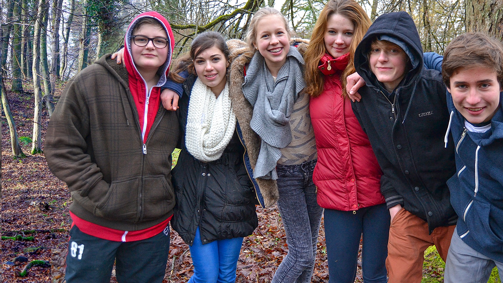 Sechs Jugendliche stehen nebeneinander und haben sich an den Schultern mit den Armen eingehakt. Sie stehen in einem kahlen Stück Wald und lächeln und lachen in die Kamera.