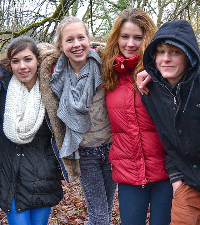 Sechs Jugendliche stehen nebeneinander und haben sich an den Schultern mit den Armen eingehakt. Sie stehen in einem kahlen Stück Wald und lächeln und lachen in die Kamera.