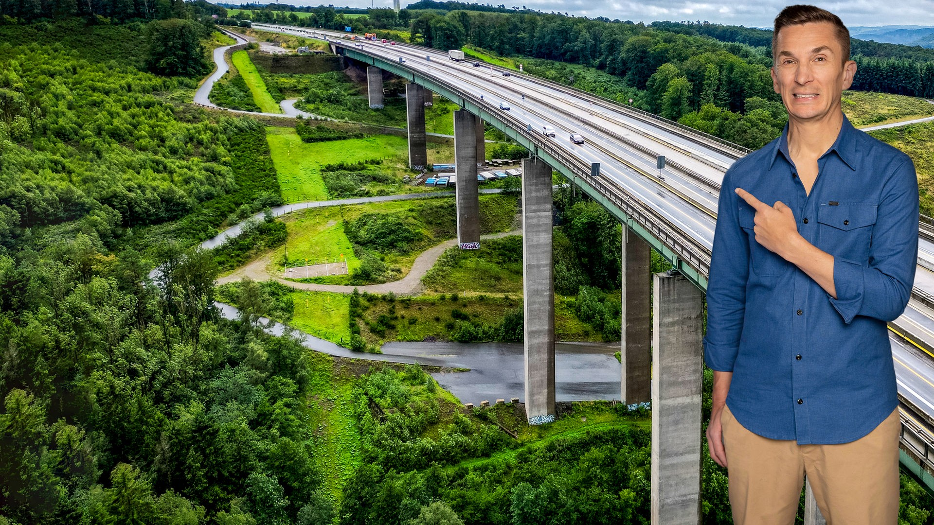 Eric Mayer steht rechts im Bild und zeigt mit dem Zeigefinger der linken Hand auf den Hintergrund, vor dem er steht. Dieser zeigt eine Autobahnbrücke, die auf vielen Pfeilern stehend sich über ein Tal schlängelt.