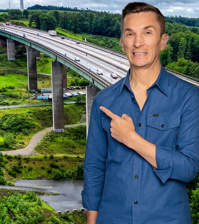 Eric Mayer steht rechts im Bild und zeigt mit dem Zeigefinger der linken Hand auf den Hintergrund, vor dem er steht. Dieser zeigt eine Autobahnbrücke, die auf vielen Pfeilern stehend sich über ein Tal schlängelt.