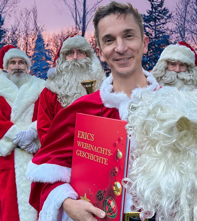 Moderator Eric Mayer steht, als Weihnachtsmann verkleidet, im Kreise weiterer Weihnachtsmänner im Freien. Er hält ein rotes Buch mit der Aufschrift "Erics Weihnachtsgeschichte" in der rechten Hand. Im Hintergrund sind verschneite Tannnen zu sehen, hinter denen die untergehende Sonne den Himmel in orangene und lila Farbverläufe taucht.