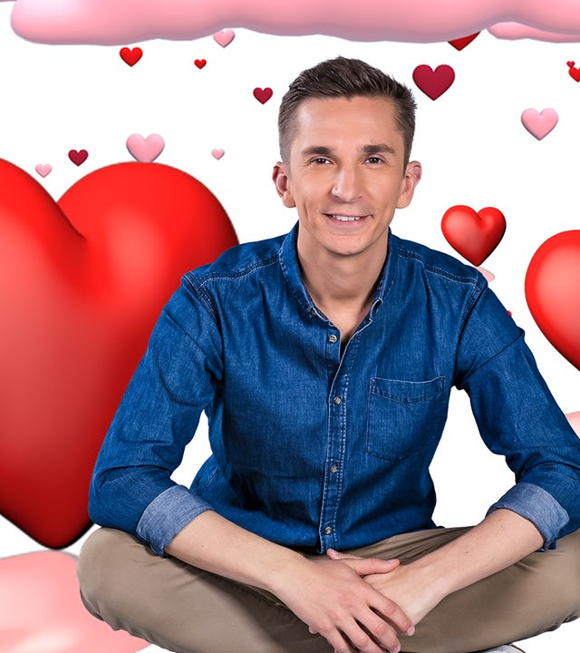 Moderator Eric Mayer sitzt im Schneidersitz und lächelt in die Kamera. Er sitzt vor einem weißen Hintergrund der gefüllt ist mit roten und rosa Herzen in verschiedenen Größen und mit weißen und rosafarbenen Wolken.
