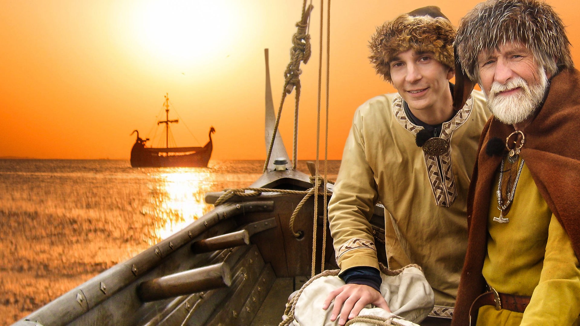 Eric Mayer rechts im Bild mit Wikinger-Kleidung und Fellmütze. Rechts neben ihm einer bärtiger, älterer Mann ebenfalls in Wikinger-Kleidung. Beide sitzen in einem Wikinger Boot. Hinter ihnen im Sonnenuntergang gleitet ein weiteres Wikinger-Boot übers Wasser.