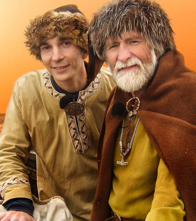 Eric Mayer rechts im Bild mit Wikinger-Kleidung und Fellmütze. Rechts neben ihm einer bärtiger, älterer Mann ebenfalls in Wikinger-Kleidung. Beide sitzen in einem Wikinger Boot. Hinter ihnen im Sonnenuntergang gleitet ein weiteres Wikinger-Boot übers Wasser.