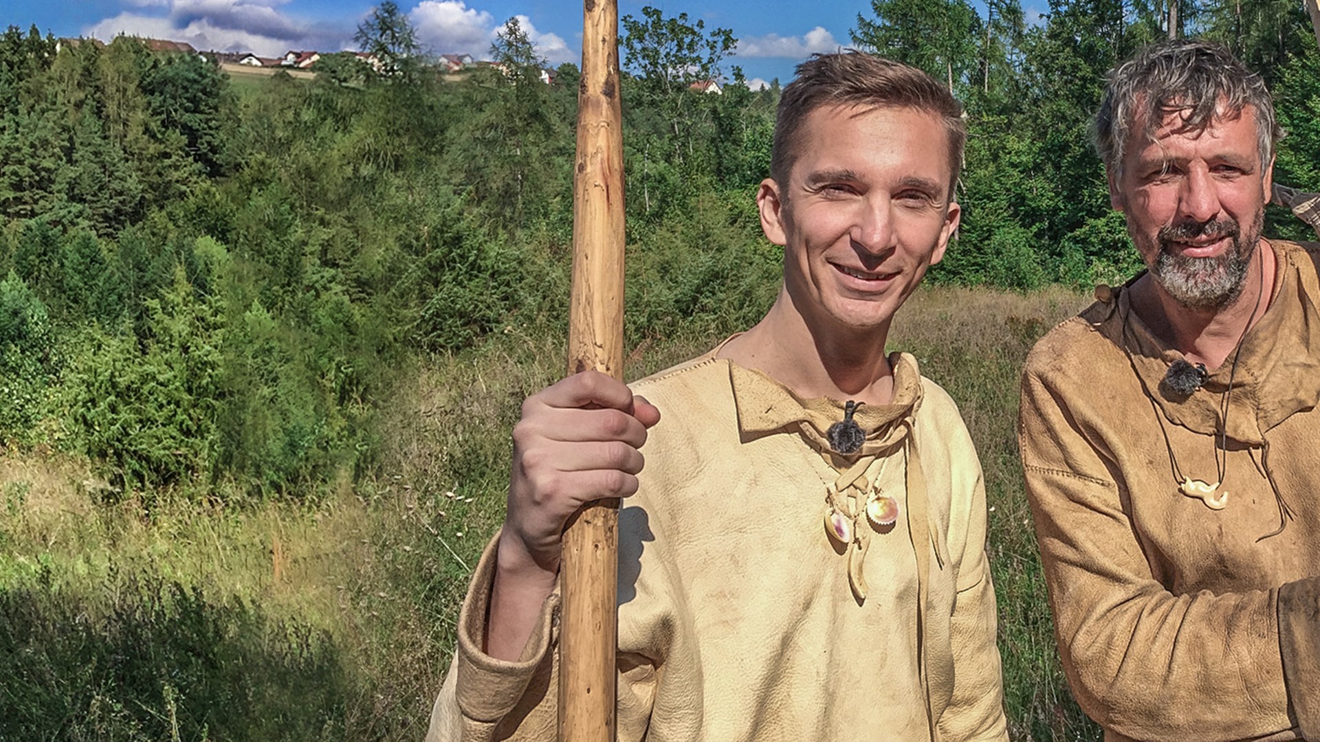 Moderator Eric Mayer steht links im Bild. Er trägt steinzeitiche Kleidung aus Leder und Lederstiefel. In der rechten Hand hält er einen riesigen Speer aus Holz. Rechts nebem ihm steht ebenso gekleidet und mit riesigen Pfeilen in der Hand ein Steinzeitexperte mit ihm auf einer Wiese.