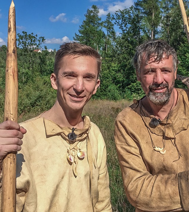 Moderator Eric Mayer steht links im Bild. Er trägt steinzeitiche Kleidung aus Leder und Lederstiefel. In der rechten Hand hält er einen riesigen Speer aus Holz. Rechts nebem ihm steht ebenso gekleidet und mit riesigen Pfeilen in der Hand ein Steinzeitexperte mit ihm auf einer Wiese.
