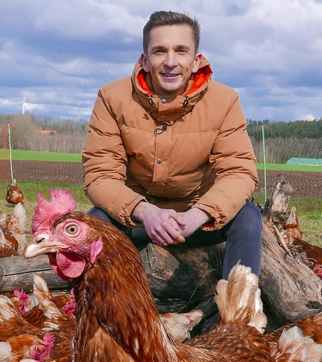 PUR+ Moderator Eric Mayer sitzt inmitten einer Schar von Hühnern.