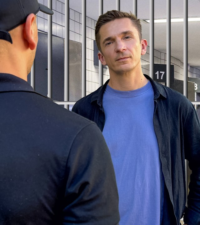 Eric steht vor einer Gittertür. Vor ihm und mit dem Rücken zur Kamera steht ein Mann und schaut ihn an. 