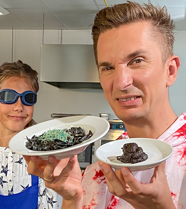 Eric hält mit angeekeltem Blick einen Teller in die Kamera, auf dem etwas Braunes, ekelig aussehendes liegt. Hinter ihm sieht man einen Jungen mit verbundenen Augen.