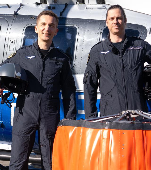 Eric Mayer steht neben Pilot Tim vor einem Hubschrauber der Polizeifliegerstaffel Nordrhein-Westfalen. Vor ihnen steht ein Außenlastbehälter, der beim Löscheinsatz unter dem Hubschrauber angehängt wird, um Wasser aus Flüssen oder Seen zu entnehmen und zu den Bränden zu transportieren.