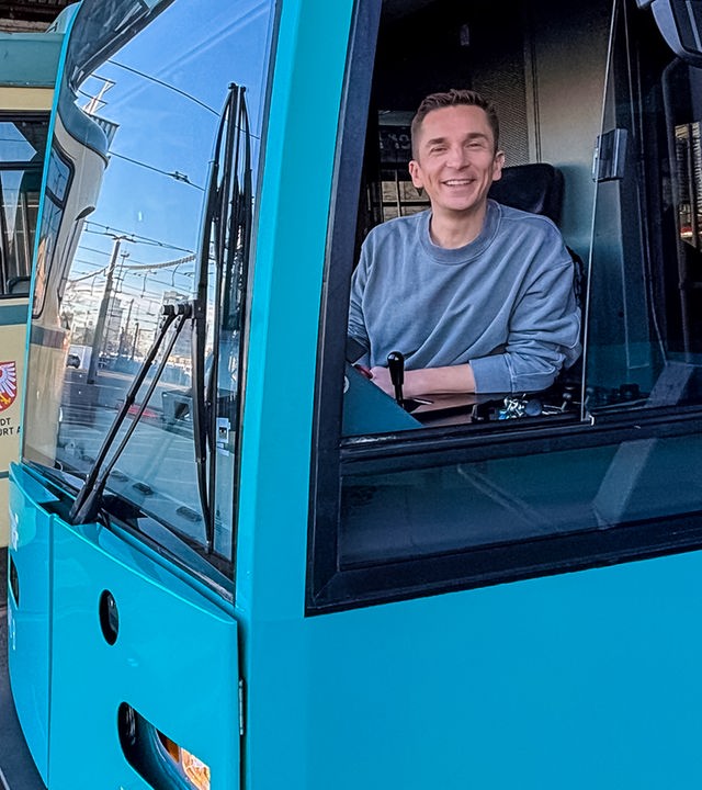 Eric sitzt im Führerhaus einer blauen Straßenbahn. Er schaut durchs offene Fenster in die Kamera und lächelt.
