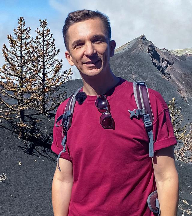 Eric schaut frontal in die Kamera. Hinter ihm sind schwarze Hügel und Berge und vertrocknet aussehende Bäume.