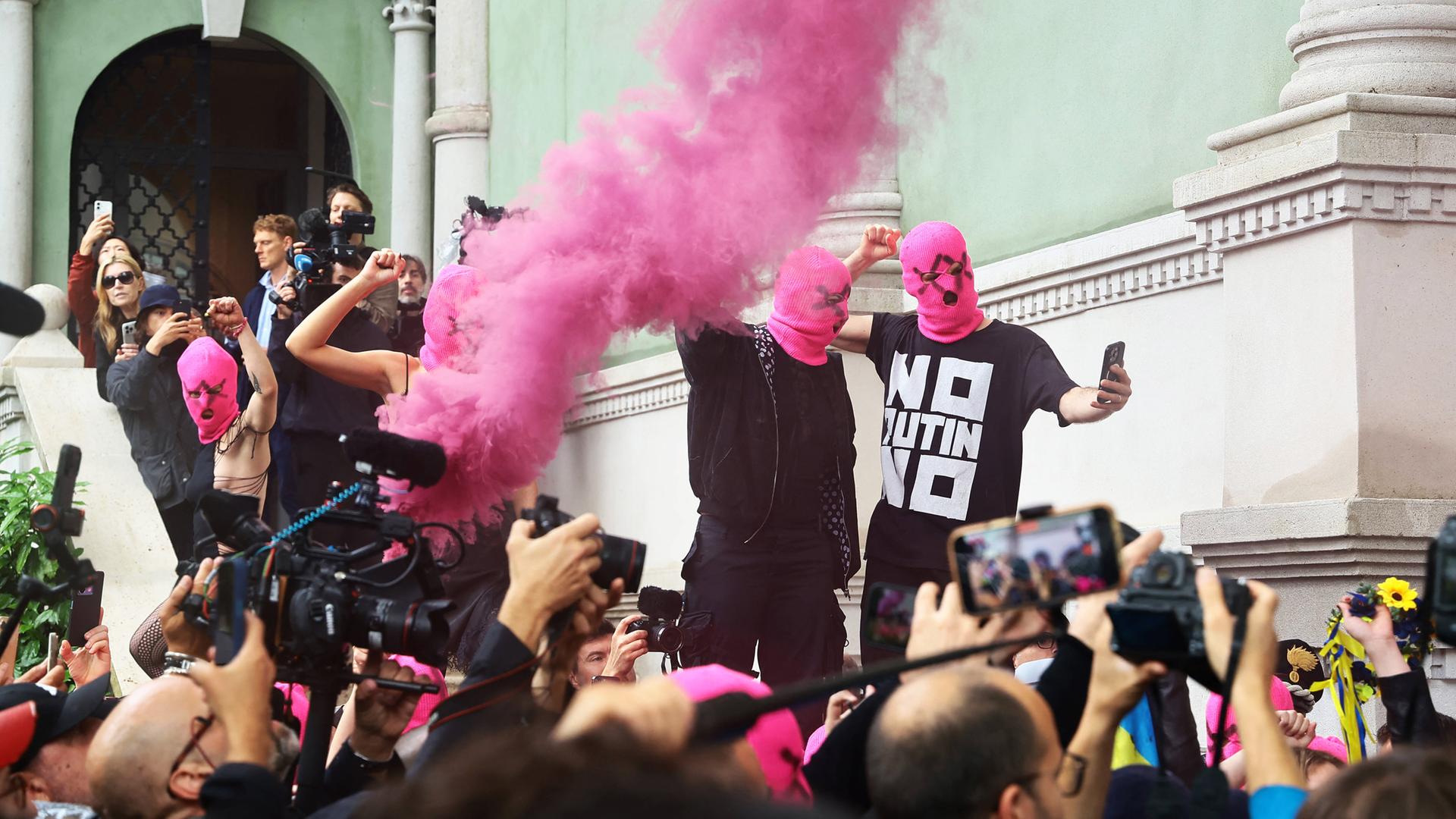 Schwarz gekleidete Menschen mit pinkfarbenen Gesichtsmasken protestieren vor einem grünen Gebäude, während eine Menge aus Reportern und Zuschauern, filmt und fotografiert.