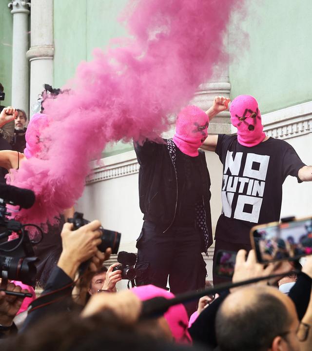 Schwarz gekleidete Menschen mit pinkfarbenen Gesichtsmasken protestieren vor einem grünen Gebäude, während eine Menge aus Reportern und Zuschauern, filmt und fotografiert.