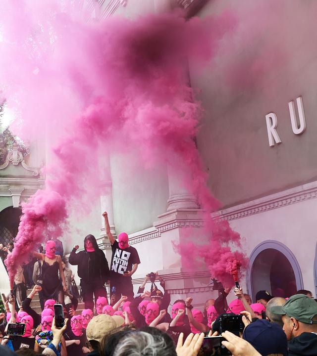 Schwarz gekleidete Menschen mit pinkfarbenen Gesichtsmasken protestieren vor einem grünen Gebäude, während eine Menge aus Reportern und Zuschauern, filmt und fotografiert.